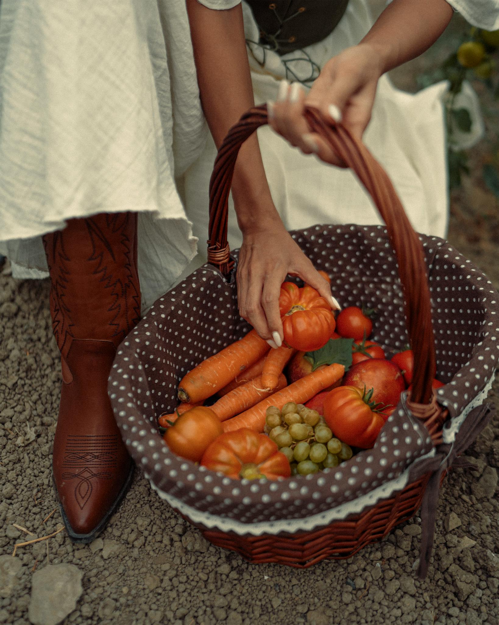 A person picking fresh produce from a basket outdoors, featuring carrots, tomatoes, and grapes.