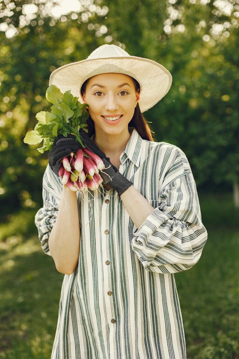 Smiling woman in hat holds freshly harvested radishes in a garden.