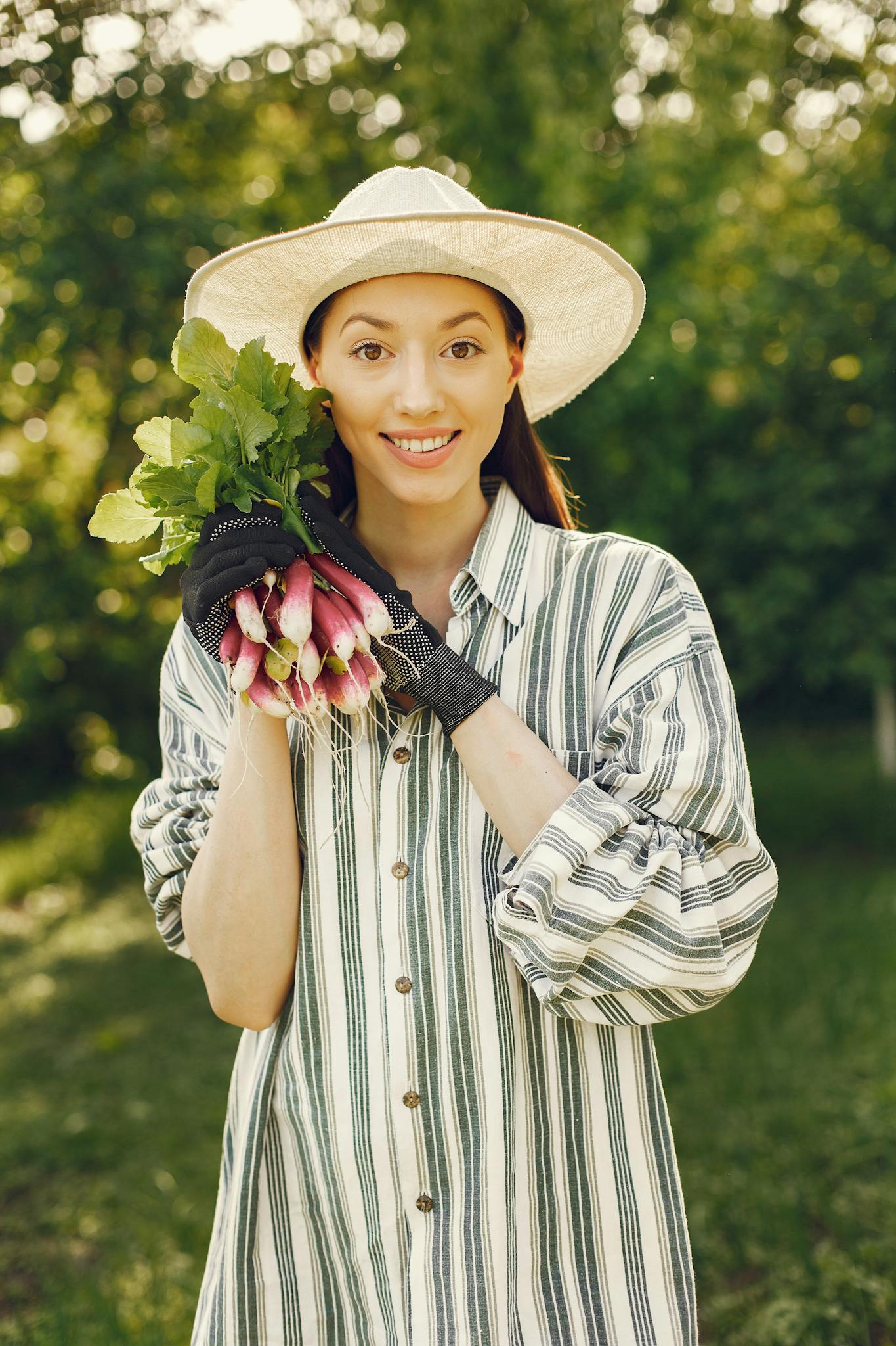Smiling woman in hat holds freshly harvested radishes in a garden.